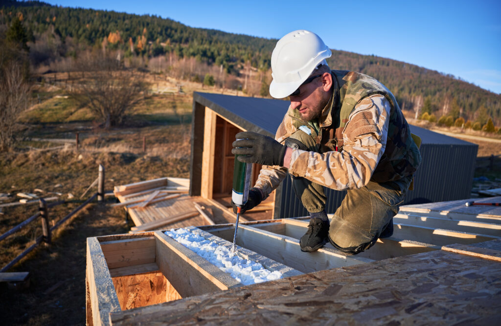 Male builder doing thermal insulation on roof of wooden frame house by polyurethane foam.